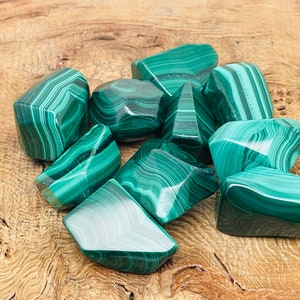 May include: A collection of polished malachite stones, showcasing a vibrant green color with distinctive banded patterns. The stones vary in size and shape, resting on a light brown wooden surface. The malachite pieces are smooth and reflect light, highlighting their natural beauty.
