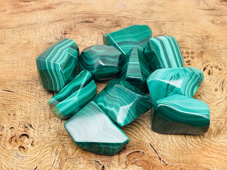 May include: A collection of polished malachite stones, showcasing a vibrant green color with distinctive banded patterns. The stones vary in size and shape, resting on a light brown wooden surface. The malachite pieces are smooth and reflect light, highlighting their natural beauty.