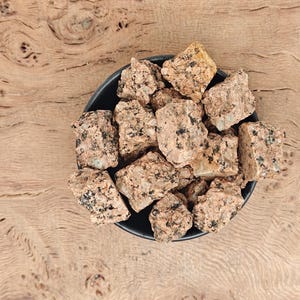 May include: A black bowl filled with rough, tan-coloured rocks with black speckles. The bowl sits on a light brown, wooden surface with a natural grain pattern. The rocks appear to be granite.
