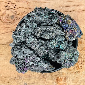 May include: A close-up of a bowl filled with iridescent black silicon carbide crystals. The crystals have a metallic sheen and are arranged in a random pattern.