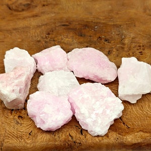 May include: A collection of rough, raw rose quartz crystals. The stones vary in size and shape, displaying a pale pink hue with some areas appearing nearly white. The crystals are arranged on a wooden surface, showcasing their natural texture.