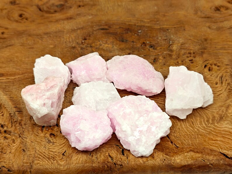 May include: A collection of rough, raw rose quartz crystals. The stones vary in size and shape, displaying a pale pink hue with some areas appearing nearly white. The crystals are arranged on a wooden surface, showcasing their natural texture.