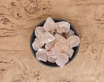 May include: A black bowl filled with raw, light pink rose quartz crystals. The crystals have a translucent appearance and varying shapes. The bowl sits on a wooden surface with a natural grain pattern.