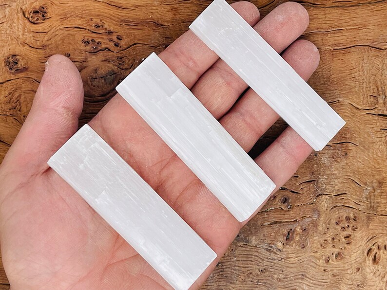 K&ouml;nnte beinhalten: Drei wei&szlig;e Selenit-St&auml;be, auch bekannt als Selenit-St&auml;be, werden in einer Hand gehalten. Die St&auml;be sind glatt und haben eine gestreifte Textur.