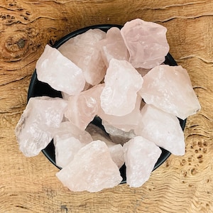 May include: A bowl filled with raw rose quartz crystals. The crystals are a light pink color and have a rough, natural texture.