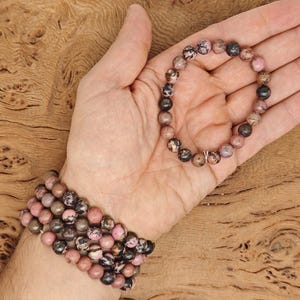 Może przedstawiać: Dwa bransoletki wykonane z różowych i czarnych koralików z kamieni szlachetnych. Bransoletki znajdują się na nadgarstku i dłoni osoby.