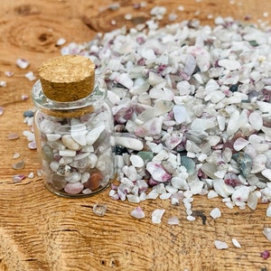 May include: A small glass jar with a cork lid filled with white, pink, and gray gemstone chips. The jar is sitting on a wooden surface with a pile of the same gemstone chips scattered around it.