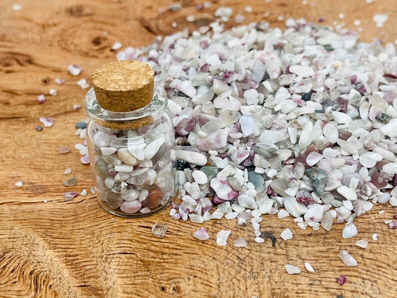 May include: A small glass jar with a cork lid filled with white, pink, and gray gemstone chips. The jar is sitting on a wooden surface with a pile of the same gemstone chips scattered around it.