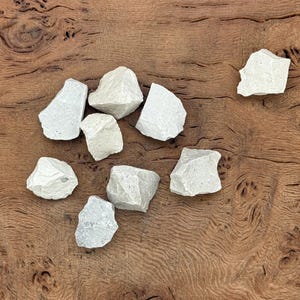 Puede incluir: Un conjunto de piedras de color gris claro y forma irregular, dispuestas sobre una superficie de madera marr&oacute;n oscura. Las piedras, de tama&ntilde;os variados y textura rugosa, sugieren un origen natural. La imagen evoca materiales org&aacute;nicos, posiblemente para artesan&iacute;a o decoraci&oacute;n.