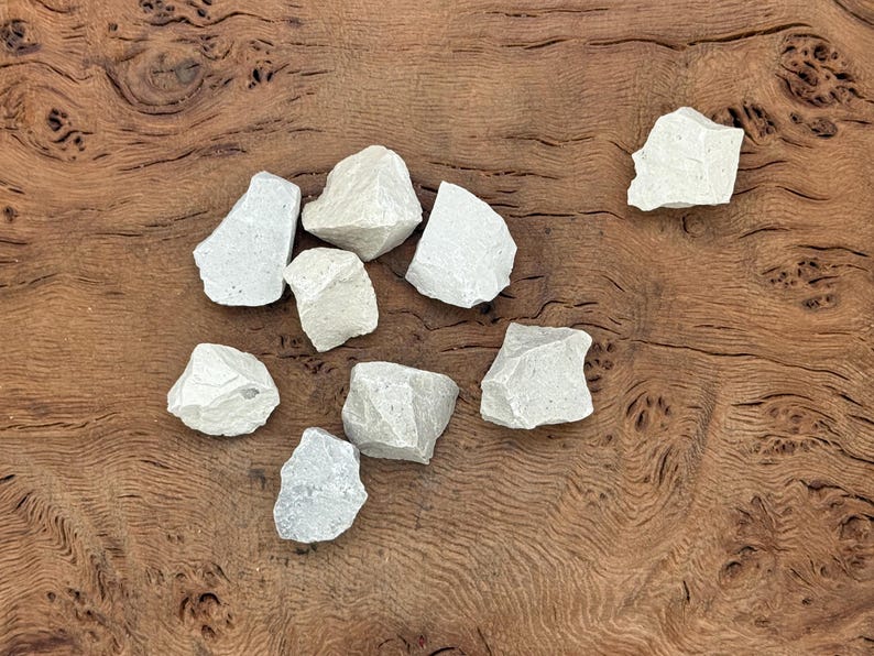 Puede incluir: Un conjunto de piedras de color gris claro y forma irregular, dispuestas sobre una superficie de madera marr&oacute;n oscura. Las piedras, de tama&ntilde;os variados y textura rugosa, sugieren un origen natural. La imagen evoca materiales org&aacute;nicos, posiblemente para artesan&iacute;a o decoraci&oacute;n.