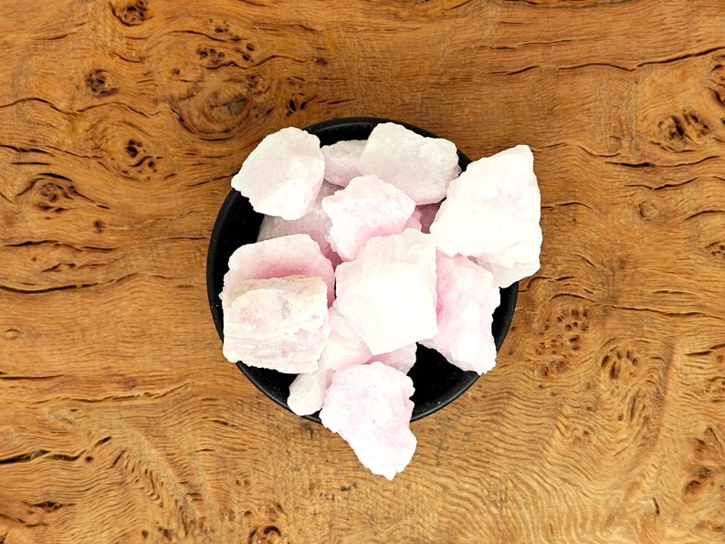 May include: A black bowl filled with rough, light pink crystals. The crystals have a natural, uneven texture and are clustered together. The bowl sits on a wooden surface with a prominent wood grain pattern.