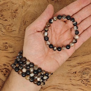 Puede incluir: Un brazalete hecho de cuentas de ágata negra y marrón. El brazalete está en la muñeca de una persona.