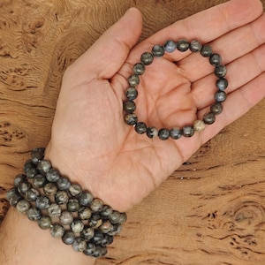 Könnte beinhalten: Zwei Armbänder aus dunkelgrauen und braunen, gefleckten Steinperlen. Die Armbänder sind auf dem Handgelenk einer Person übereinander gelegt.