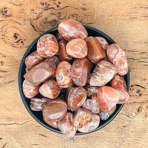 May include: A bowl of carnelian tumbled stones. The stones are a mix of light and dark orange with white patterns. The bowl is black and the stones are on a wooden surface.