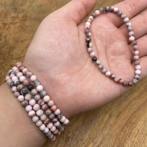 Puede incluir: Varios brazaletes de cuentas en tonos rosa, gris y marrón. Un brazalete se sostiene en una mano, mientras que otros están apilados en una muñeca. Las pequeñas cuentas redondas crean una estética delicada y femenina.