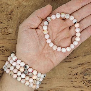 May include: A collection of pink gemstone bracelets. One bracelet is held in an open hand, while several others are stacked on a wrist. The beads have a mix of pink, white, and black speckles. The background is a wooden surface.