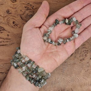 May include: Two bracelets made of green and clear chip stones. The bracelets are stacked on a person's wrist.