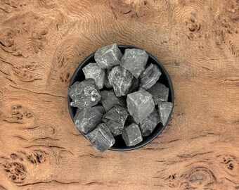 May include: A black bowl filled with rough, gray stones. The stones have a textured appearance and varying shapes. The bowl is set against a warm, wooden background, creating a contrast in color and texture.