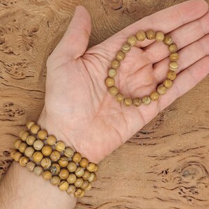 May include: A close-up of a hand wearing a bracelet made of multiple strands of light brown beads. The bracelet is made of natural stone beads with a slightly marbled pattern.