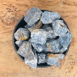 Könnte beinhalten: Eine Schale gefüllt mit rauen, grauen und blauen Trommelsteinen. Die Steine haben einen leicht metallischen Glanz und sind in einem zufälligen Muster angeordnet.