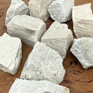 Puede incluir: Un conjunto de piedras de color gris claro, de forma irregular y con superficie rugosa y texturizada. Las piedras son de diferentes tama&ntilde;os y est&aacute;n dispuestas sobre una superficie de madera, creando una est&eacute;tica natural y terrosa. Ideal para manualidades o decoraci&oacute;n.