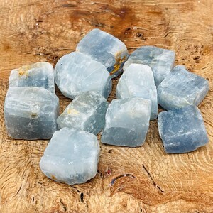 May include: A collection of ten blue calcite crystal chunks, with a rough, natural texture. The crystals are various sizes and shapes, and are arranged on a wooden surface.