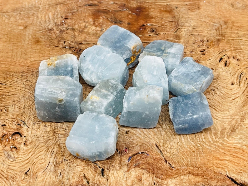 May include: A collection of ten blue calcite crystal chunks, with a rough, natural texture. The crystals are various sizes and shapes, and are arranged on a wooden surface.