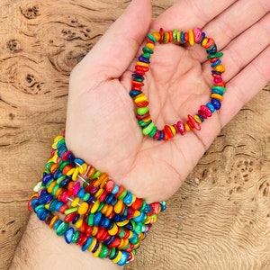 Könnte beinhalten: Ein farbenfrohes Armband aus kleinen, flachen Holzperlen. Das Armband wird an einem Handgelenk und einer Hand gezeigt. Die Perlen sind in einem Regenbogenmuster angeordnet.