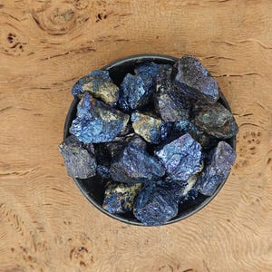 May include: A black bowl filled with iridescent blue and grey rough-cut stones. The stones have a metallic sheen and varying textures. The bowl sits on a wooden surface with a natural grain pattern.