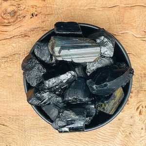 Puede incluir: Un cuenco negro lleno de cristales de turmalina negra. Los cristales son ásperos y tienen un aspecto natural y sin pulir.