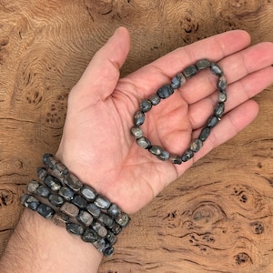 Könnte beinhalten: Ein Armband aus dunkelgrauen und schwarzen Edelsteinperlen. Das Armband besteht aus mehreren Perlensträngen und wird an einem Handgelenk getragen.