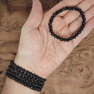 May include: Two black beaded bracelets, one is a single strand and the other is a multi-strand bracelet. The bracelets are on a person's wrist.