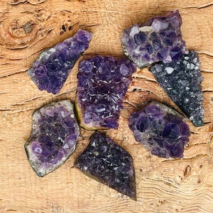 Puede incluir: Seis grupos de cristales de amatista de varios tamaños y formas, dispuestos sobre una superficie de madera. Los cristales son de color púrpura intenso y tienen una textura natural y rugosa.