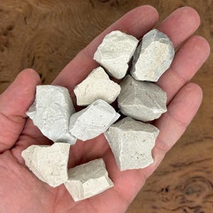 Puede incluir: Un pu&ntilde;ado de rocas de color gris claro, de forma irregular. Las rocas tienen una superficie rugosa y texturizada y diferentes tonos de gris y blanquecino. Las rocas se sostienen en la mano de una persona, con un fondo de madera.