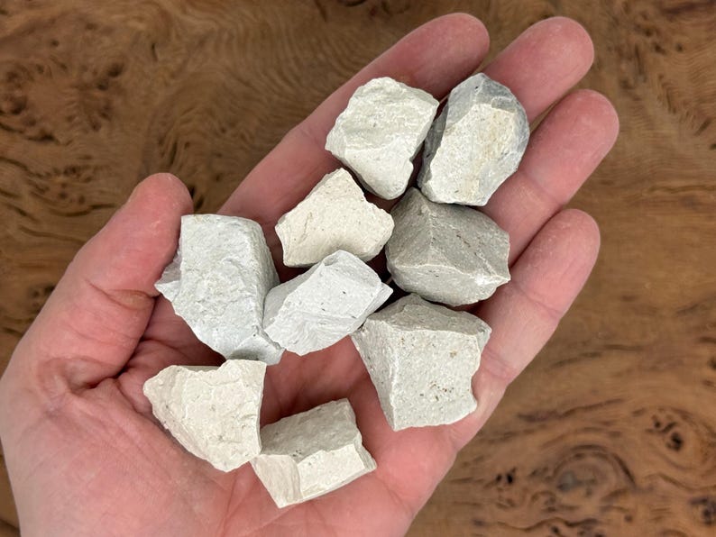 Puede incluir: Un pu&ntilde;ado de rocas de color gris claro, de forma irregular. Las rocas tienen una superficie rugosa y texturizada y diferentes tonos de gris y blanquecino. Las rocas se sostienen en la mano de una persona, con un fondo de madera.