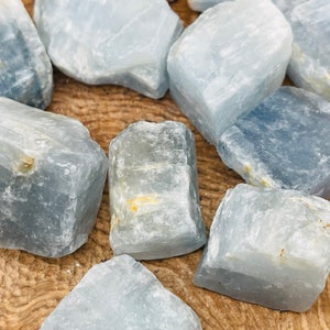 May include: A collection of eight light blue celestite crystal roughs with a natural, raw, and unpolished finish. The crystals are various sizes and shapes, with some showing a slight golden hue.