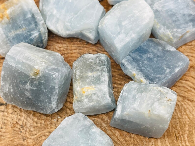 May include: A collection of eight light blue celestite crystal roughs with a natural, raw, and unpolished finish. The crystals are various sizes and shapes, with some showing a slight golden hue.