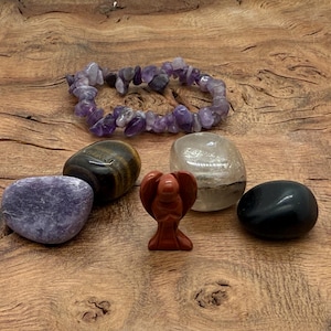 May include: A collection of gemstones and a bracelet on a wooden surface. The gemstones include a purple amethyst bracelet, a tiger's eye stone, a clear quartz stone, a red carved angel, a black obsidian stone, and a purple stone.