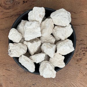 May include: A black bowl filled with numerous off-white, irregularly shaped rocks. The rocks have a rough, textured surface and are clustered together. The bowl is set against a wooden background with a natural grain pattern.