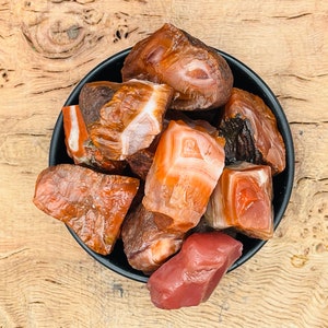 Könnte beinhalten: Eine Schale mit rohen Karneol-Steinen. Die Steine sind in einer Mischung aus orange, rot und braun gefärbt. Die Schale ist schwarz und die Steine liegen auf einer Holzoberfläche.