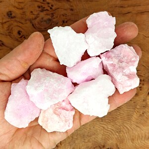 May include: A collection of raw, rough-cut rose quartz crystals in varying shades of pink and white. The crystals are held in a hand, showcasing their natural, unpolished texture. The background is a wooden surface.