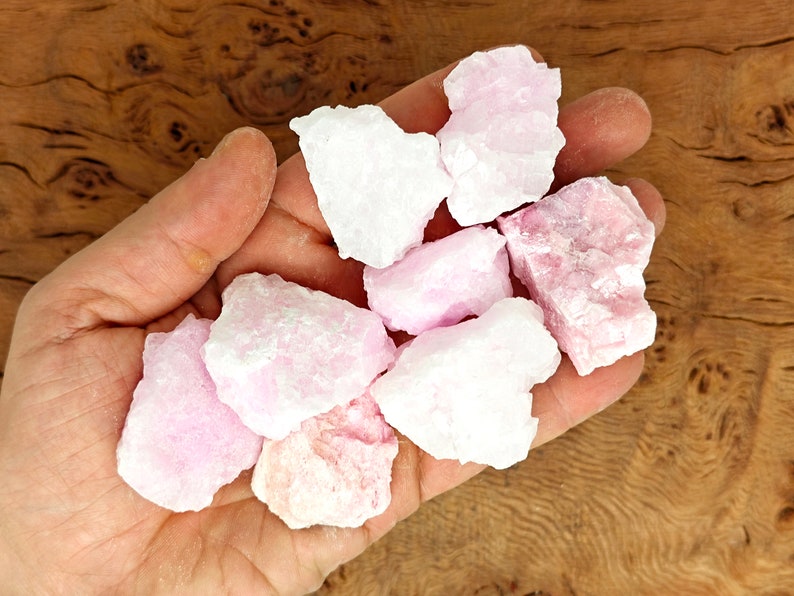 May include: A collection of raw, rough-cut rose quartz crystals in varying shades of pink and white. The crystals are held in a hand, showcasing their natural, unpolished texture. The background is a wooden surface.