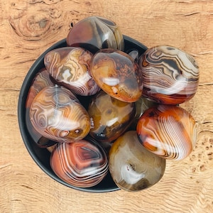 Puede incluir: Una colección de piedras de ágata bandeada pulidas en un cuenco negro. Las piedras son una mezcla de colores marrón, blanco y negro con intrincados patrones.