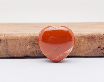 May include: A polished, heart-shaped stone in shades of red and orange, with subtle white and gold veining. The gemstone is smooth and reflective, set against a light wood surface. A decorative item.