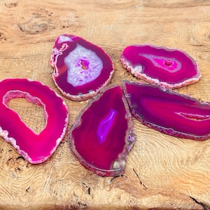 Pode incluir: Cinco fatias de ágata polidas em tons de rosa e roxo, com padrões de bandas únicos. As pedras preciosas são exibidas em uma superfície de madeira natural, mostrando suas cores vibrantes e sua beleza natural. Ideal para decoração de casa ou artesanato.