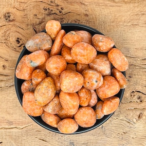 May include: A bowl filled with orange carnelian tumbled stones. The stones are smooth and polished, with a variety of shapes and sizes.