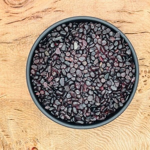 May include: A black bowl filled with small, dark red, polished stones. The bowl is sitting on a light brown wooden surface.