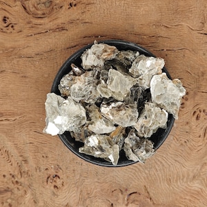May include: A bowl filled with a variety of light grey and silver mica crystals. The crystals are irregularly shaped and have a shimmery, iridescent quality.