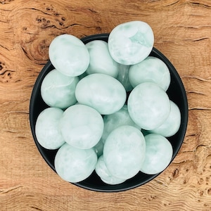 Puede incluir: Un cuenco negro lleno de piedras lisas de color verde pálido. Las piedras tienen forma ovalada y redondeada y un aspecto ligeramente translúcido. El cuenco está sobre una superficie de madera con un veteado natural.