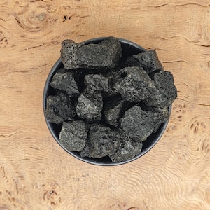 Pode incluir: Uma tigela preta cheia de pedras de lava cinza escuro. A tigela está sobre uma superfície de madeira marrom claro.